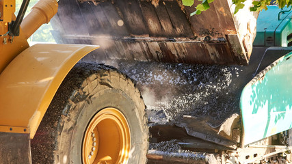 Bucket tractor pours hot asphalt into a grader, construction of a new road, material for road construction close-up.