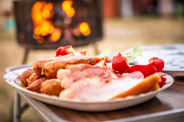 Close up of raw bacon, sausages and paprika on colorful tray, prepared for barbecue. With glowing grill in the background