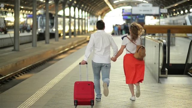 Running Couple With A Suitcase In A Train Station. Woman Holding Passports In Her Hands.