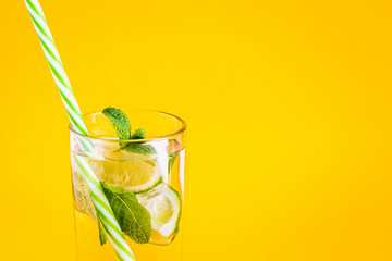 Fresh summer lemonade with water, lime and mint in a glass tumbler with a straw on a yellow background. Cold, refreshing summer cocktail close up. Fresh drink with copy space