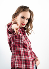 Beautiful emotional girl model with red lips make-up, wearing a casual plaid shirt and jeans points her finger at the viewer with a raised eyebrow. Isolated on white