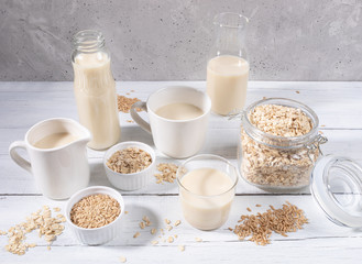 Set of different containers with oat milk, oat seeds and flakes on white wooden table on concrete background. 
