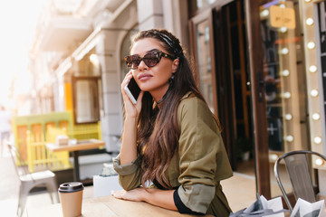 Elegant girl with long dark hair calling someone while resting in outdoor cafe with cup of coffee. Outdoor photo of serious fashionista woman in green coat talking on phone on blur city background.