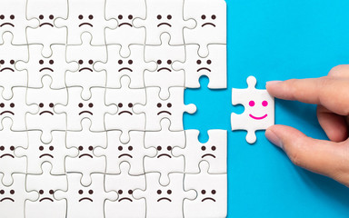 Hand of a businessman chooses a smiley face on piece of white jigsaw puzzle on blue background, The...