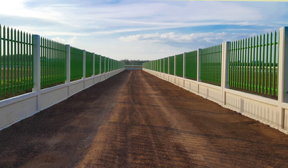 The gravel road lies between the white cement fence with green iron bars under the blue sky.