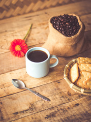 Black coffee In the cup sky blue color have roasted coffee beans are In a cloth bag sack butter cracker In the basket weave bamboo, still have red flowers and spoon all placed on a wooden table.