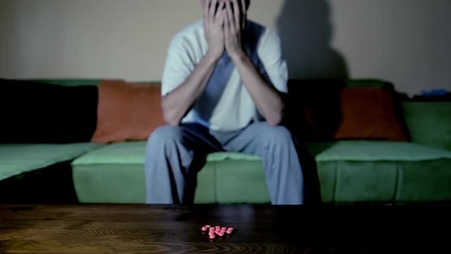 Depressed man suffering from suicidal depression want to commit suicide by taking strong medicament drugs and pills pain killers while he sitting in his dark room selective focus moody dramatic look