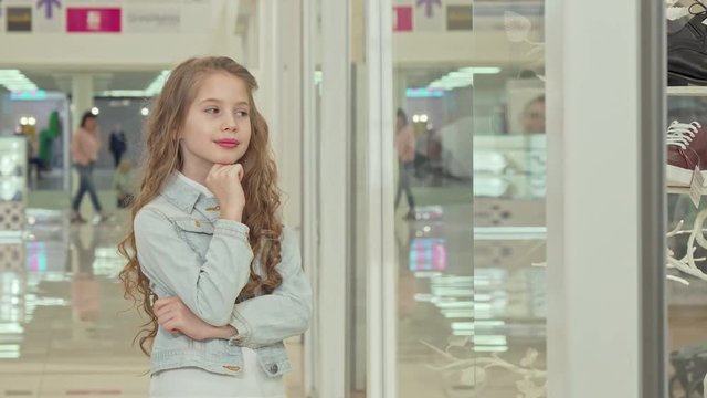 Cute little girl smiling to the camera, examining fashion store display. Adorable beautiful girl looking at clothing boutique showcase. Happy lovely girl at the shopping mall