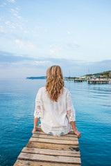 A girl sits on a pier in the morning enjoying the azure sea.
