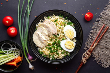 Asian ramen noodles with chicken, vegetables and egg in black bowl on black background. Top view.