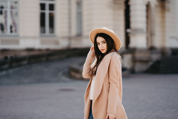 Fototapeta premium Beautiful young woman wearing long beige coat walking through the city streets. Trendy casual outfit. Street fashion.