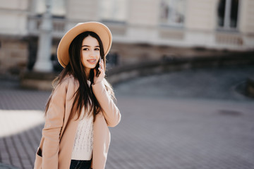 Beautiful Caucasian woman talking by phone with friend while standing on sunny street. Brunette wearing beige coat, looking away, has important conversation. Communication concept.