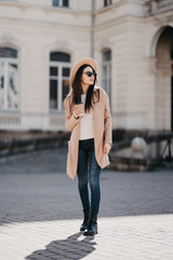 Fashion outdoor photo of beautiful girl with dark hair in elegant beige coat walking by street with a cup of coffee