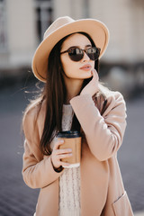 Fototapeta premium Portrait of young woman wearing coat and eyeglasses drinking takeaway coffee while walking through city street