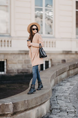 Young woman wearing coat and black hat walking in city street and drinking take away coffee in...