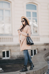 Outdoor shot of attractive young woman wearing coat in sunny city street and drinking take away coffee in paper cup, keeps hand on her sunglasses, going to office after lunch breack. People concept.