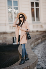 Young woman wearing coat and black hat walking in city street and drinking take away coffee in...