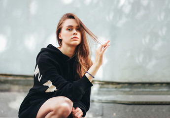 Fototapeta premium Candid portrait of young beautiful long hair unhappy girl fashion model hipster in black hoodie on wall background
