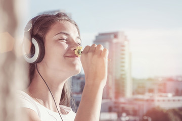 teenager girl listening to music on headphones on balcony of urban house, smelling flower with...