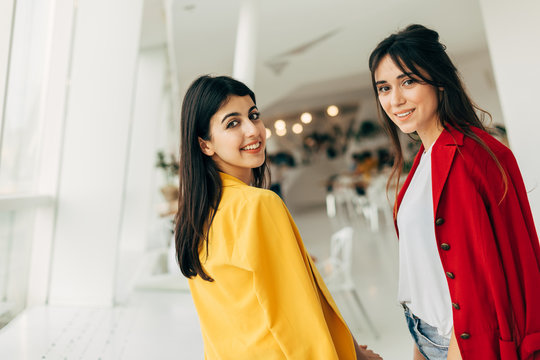 Beautiful cheerful attractive young women look back and posing on camera together. Wear red and yellow jacket. Stand in white cafe room. Stylish models together.