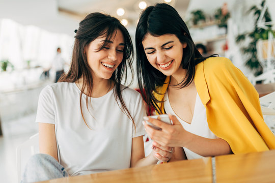 Cheerful happy nice young businesswomen in coworking. Sitting together in white room and laughing. Girl on right side holding phone in hands. Casual stylish workers.