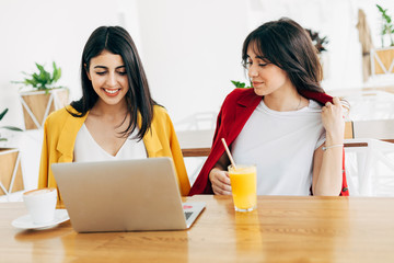 Beautiful attractive young women in coworking space. Working together. Stylish businesswomen in cafe. Model on left typing on keyboard. Woman on right drink orange juice. Sit on table.