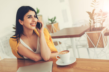 Nice cheerful attractive young woman sitting at table and talking on phone. Smiling. Cup of coffee and laptop on table. Room with white walls or coworking.
