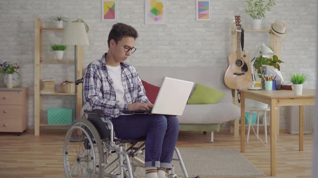 portrait young Asian man in stylish glasses disabled in a wheelchair with a laptop in the living room of the house
