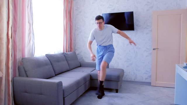 Funny nerd man is making squats and slopes to the legs and touches the legs with his hands, exercise at home in living room. Funny pulls up shorts before exercise. Front view.
