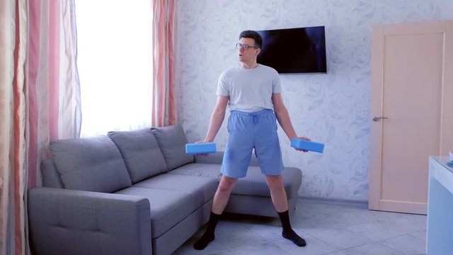 Playful nerd man in glasses and shorts is doing exercises for hand biceps with yoga blocks instead of dumbbells with efforts at home. Sport humor concept.