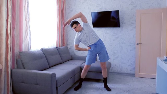 Funny nerd man in glasses and blue shorts is doing complex of fitness exercises at home. Sport humor concept.
