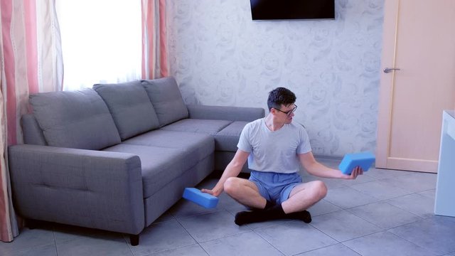 Weak nerd man in glasses is doing exercises for hands with yoga blocks instead of dumbbells with efforts sitting on the floor at home. Sport humor concept.