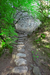 A stone pathway leading throughan archway of bright green tree leaves to a stone wall with opening and dappled sunlight.