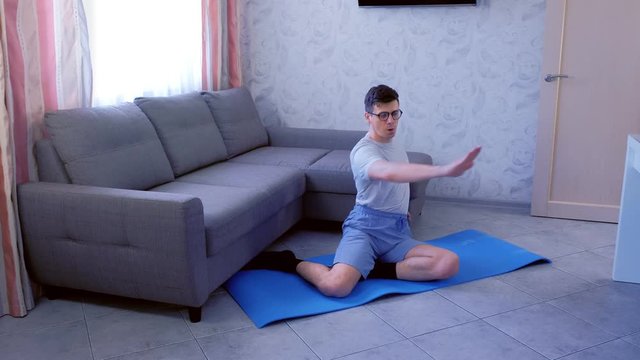 Funny nerd man in glasses is doing shaping hands exercises sitting on mat at home. Sport humor concept.