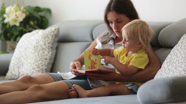 Sweet toddler boy and his mother, sitting on a couch, eating cherries and looking at picture book, enjoying healthy meal