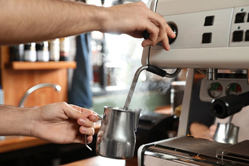 Fototapeta na wymiar Barista steaming milk in metal jug with coffee machine wand at bar counter, closeup