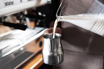 Barista pouring milk into metal pitcher indoors, closeup with space for text. Coffee making