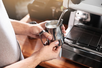 Barista frothing milk in metal pitcher with coffee machine wand at bar counter, closeup