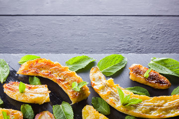 Halves of grilled bananas on a black kitchen board and shale stone. Baked banana dessert with mint on black background. Asian healthy food. Top view © Lazartivan