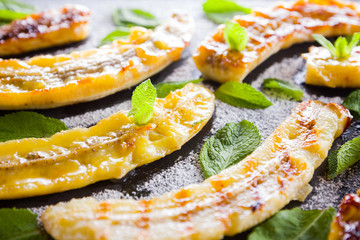Halves of grilled bananas with powdered sugar on a black kitchen board and shale stone. Baked banana dessert with mint on black background table. Close up view