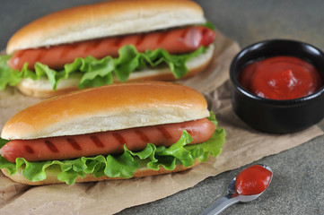 Two hot dogs on craft paper. A cup with ketchup sauce and a spoon with a sauce. Close-up. Dark background.