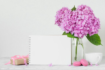 Modern still life with pink hydrangea flowers, blank paper card, coffee cup and gift box on white wooden tabl