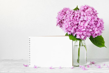 Obraz premium Modern still life with a beautiful pink hydrangea flowers and blank paper card on white wooden table