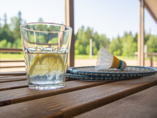 Refreshing lemonade with a slice of lemon in the foreground. Two badminton rackets and shuttlecock on wooden table. Lush green foliage. Wooden country furniture on the terrace of a country house.