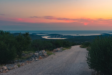 Landscape view of a gravel road and the Croatian coastline with scattered islands and a village with a marina with sailing boats in the distance during a beautiful sunset in summer, Dalmatia, Croatia