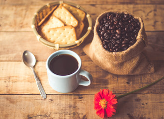 Black coffee In the cup sky blue color have roasted coffee beans are In a cloth bag sack butter cracker In the basket weave bamboo, still have red flowers and spoon all placed on a wooden table.