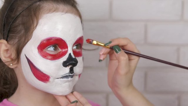 The child is painted face for Halloween. A cute little girl gets a scary face for Halloween.