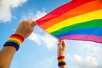 Hands with rainbow color wristbands waving gay pride flag backlit in the wind against a soft blue...