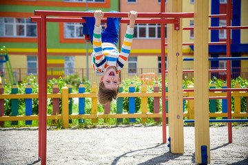 Happy child boy hanging upside down on bar, playground in city, outdoor activities.