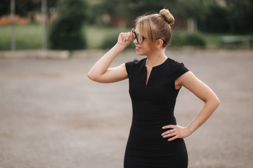 Young attractive female techer in black dress with sunglasses standing outside. Background of school